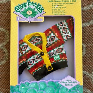 May include: A Cabbage Patch Kids Deluxe Fashions long-sleeve cardigan in a red, white, and green geometric knit pattern with a yellow collar, black cuffs, and two black buttons, packaged in a yellow and purple cardboard box with product text.