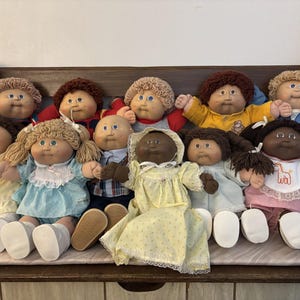 May include: A collection of Cabbage Patch Kids dolls, each with unique features. The dolls have various hair colors and styles, and wear different outfits, including dresses and overalls. Some dolls have blue eyes, and others have brown eyes.