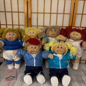 May include: A collection of six Cabbage Patch Kids dolls with yarn hair, soft bodies, and vinyl faces. The dolls wear various outfits including blue jackets, white dresses, and denim pants. A pink birth certificate document is placed in the foreground.