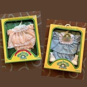 May include: Two Cabbage Patch Kids baby clothes sets in yellow boxes. One set includes a peach-colored dress with white lace trim. The other set features a light blue dress with a white collar, matching bloomers, and a pair of white shoes.