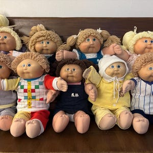 May include: A collection of nine Cabbage Patch Kids dolls with yarn hair, soft fabric bodies, and vinyl faces. The dolls are dressed in various outfits, including a white shirt with a colorful grid pattern, a yellow sleeper, and a blue and white striped baseball jersey.