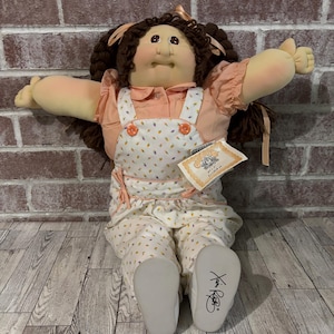 May include: A vintage Cabbage Patch Kid doll with brown hair styled in pigtails. The doll wears a peach-colored blouse, a white and patterned overall, and matching pants. The doll is seated with arms outstretched. The doll's feet are white and signed.