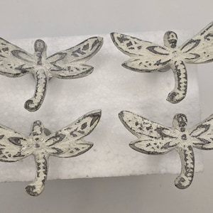 May include: Four white metal dragonfly drawer pulls with black painted details. The dragonfly design is detailed and intricate.