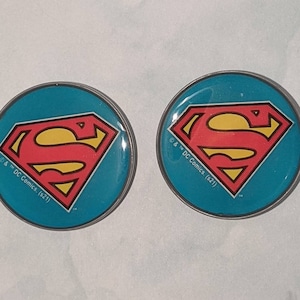 May include: Two round Superman logo pins. Each pin features the iconic red and yellow Superman emblem on a teal background, with a silver border. The pins have the text "& DC Comics. (s21)" around the edge.