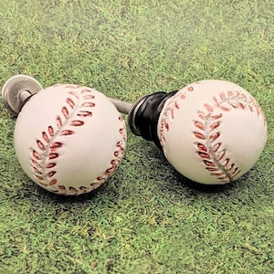 May include: Two white baseball-themed cabinet knobs with red stitching. The knobs are on a green artificial grass background.