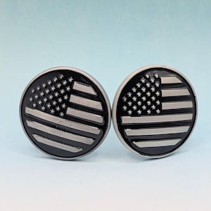 May include: Pair of round, silver-toned cufflinks with a black enamel American flag design. The flag features white stripes and a field of white stars on a black background. The cufflinks are set against a light blue and white gradient background.