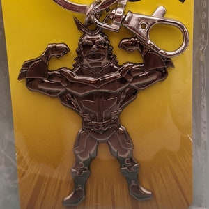 May include: A silver keychain featuring a muscular character from the anime My Hero Academia. The character is in a heroic pose with arms raised. The keychain is packaged on a yellow and black card with the text "My Hero Academia" and "Keyring".