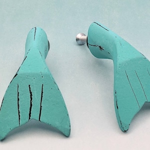 May include: A pair of turquoise blue mermaid tail earrings with black lines. The earrings are made of a lightweight material and have a silver-toned post.