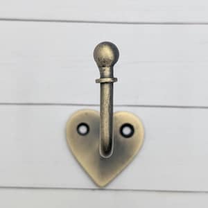 May include: A heart-shaped, antique brass wall hook. The hook has a rounded top and a vertical stem, attached to a heart-shaped base with two mounting holes. The finish is a warm, aged brass tone, suitable for hanging items.