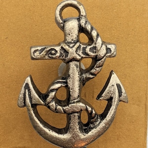 May include: A silver-toned metal anchor with a rope design. The anchor is on a brown cardboard background.