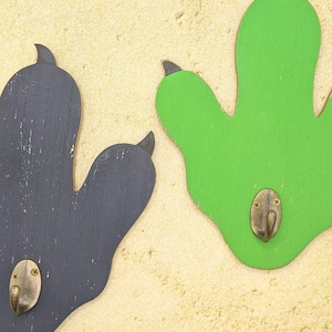 May include: Two wooden dinosaur footprint hooks, one painted black and one painted green, mounted on a beige wall.