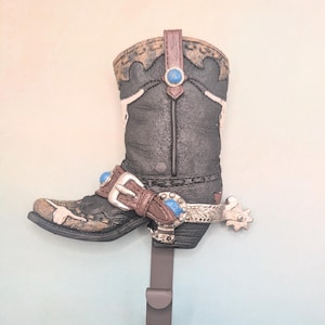 May include: A brown cowboy boot with a spur and a blue stone on the top. The boot is attached to a metal hook.