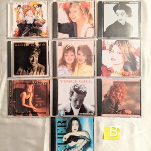 May include: A collection of nine compact discs (CDs) featuring various artists, including Shania Twain, Vince Gill, and Cher. The CDs are arranged in a grid pattern on a white surface. The CDs are all in black cases with colorful artwork.