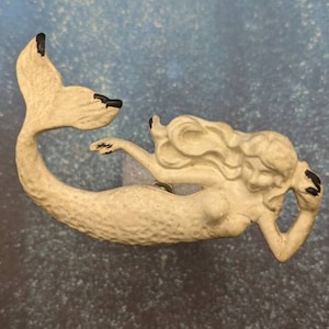 May include: White ceramic mermaid figurine with a black tail fin. The mermaid is laying on her side with her tail curled up behind her. She has long flowing hair and is holding her hand to her cheek.