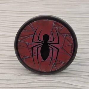 May include: A round, black phone grip with a red and black Spider-Man logo. The logo is cracked and has a web design. The text "MARVEL" is printed on the side of the grip.