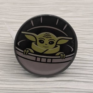 May include: A round, black and gray pin with a cartoon image of a green character from the Star Wars franchise, "The Mandalorian", in a floating pod. The character is looking out of the pod.