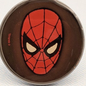 May include: A round, silver-colored pin with a red and black Spider-Man mask image. The pin has the text "©MARVEL" on the side.