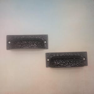 May include: Two dark gray, rectangular drawer pulls with intricate, geometric designs. Each pull has two holes for mounting and a curved handle. The pulls are made of metal and have a slightly textured surface.