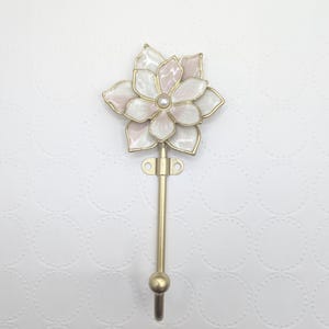 May include: A decorative wall hook featuring a flower design. The flower has white and pink petals with a gold-colored outline and a pearl-like center. The hook is gold-colored and has a rounded end.
