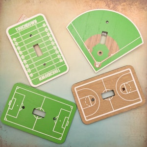 May include: Set of three light switch covers featuring a green football field, a green baseball field, and a brown basketball court. Each cover has a white outline of the field and a white light switch.