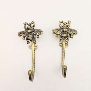 May include: Two gold-toned metal hooks shaped like bees. Each hook has a detailed bee design with wings, legs, and a stinger. The hooks are attached to a wall.