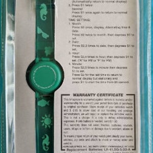 Peut inclure: Une montre Barbie pour filles, de couleur verte et blanche, avec un affichage num&eacute;rique. La montre pr&eacute;sente divers motifs sur le th&egrave;me de la mer, notamment un soleil, un hippocampe et des poissons. La montre est emball&eacute;e dans un bo&icirc;tier en plastique transparent avec un fond blanc et un certificat de garantie.