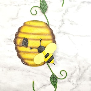 May include: A metal beehive with a bee hanging off the side. The beehive is yellow and brown with a textured surface. The bee is yellow and black with a green leaf on the side. The beehive is attached to a green metal stem with two leaves.