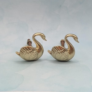 May include: Two gold-colored swan-shaped cabinet knobs. The knobs are detailed with feathers and have a classic design. The knobs are mounted on brown stems. The background is a light blue color.