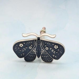May include: A decorative bottle stopper featuring a black moth design with white floral accents. The moth has detailed wing patterns and a silver-colored handle. The stopper is designed to fit into a bottle.