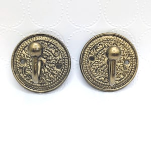 May include: Two antique brass-colored wall hooks. Each hook has a round, ornate backplate with a decorative pattern and a small hook. The hooks are mounted on a white surface with a subtle dotted pattern.