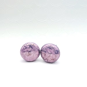 May include: Two round, marbled earrings in shades of lavender and purple. The earrings have a glossy finish and are set against a white background with a subtle, textured pattern.