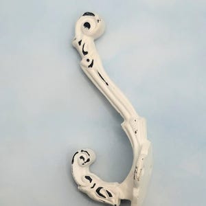 May include: White decorative wall hook with an ornate, scroll-like design. The hook has a distressed finish with black accents, adding a vintage aesthetic. The hook is made of metal and is designed for hanging items.