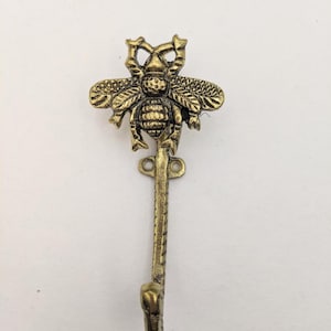 Peut inclure: Un crochet en m&eacute;tal dor&eacute; en forme d'abeille avec un design d&eacute;coratif. Le crochet est fix&eacute; &agrave; une longue tige incurv&eacute;e.