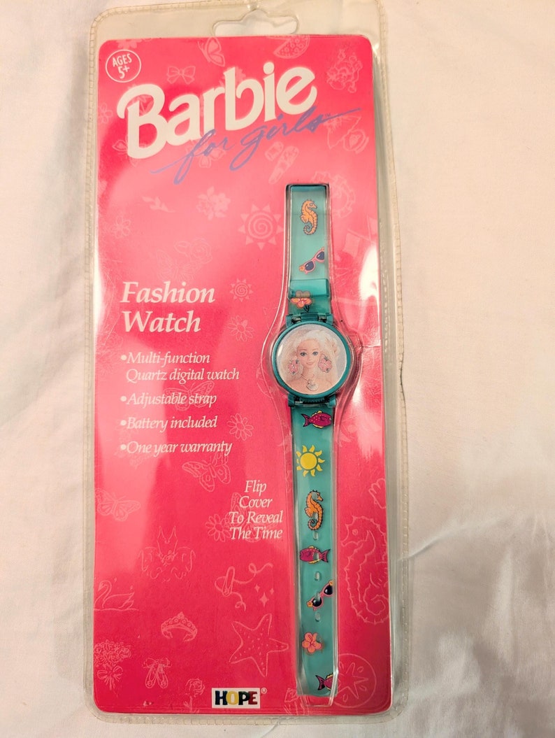 Peut inclure: Une montre Barbie bleu turquoise avec un couvercle rabattable pour r&eacute;v&eacute;ler l'heure. Le bracelet de la montre a un hippocampe, un poisson, des lunettes de soleil et un motif soleil. L'emballage est rose avec les mots "Barbie for girls" et "Fashion Watch".