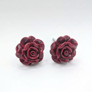 May include: Two burgundy rose-shaped cabinet knobs. The knobs are made of a glossy material and feature detailed petal designs. The knobs are mounted on silver-colored posts and are set against a white background.