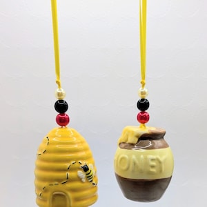 May include: Two ceramic hanging ornaments featuring a yellow beehive with a bee and a brown and yellow honey pot with the word HONEY embossed on the front. Each ornament hangs from a yellow ribbon accented with a white, black, and red bead.