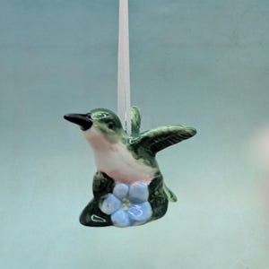 May include: A ceramic hummingbird ornament with green, white, and black details. The bird is perched on a small base with blue flowers. A white string is attached for hanging.