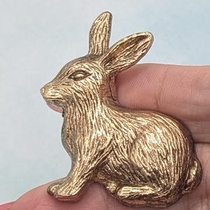 May include: A gold-colored, textured rabbit figurine in a seated position. The bunny has detailed fur and upright ears. The figurine is likely made of metal and is a decorative item.
