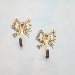 May include: Two decorative brass bow-shaped wall hooks. Each hook features a detailed bow design with a curved hook below. The hooks are a warm gold color and are mounted on a light blue and white background. Ideal for hanging items in a bedroom or bathroom.