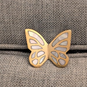 May include: A gold-colored butterfly-shaped decorative object with inlaid mother-of-pearl sections. The butterfly is resting on a gray, textured surface. The wings have a detailed design, and the overall aesthetic is elegant and decorative.