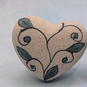 May include: Heart-shaped stone with a beige, textured surface. Black swirling vines and green leaves are painted on the stone. The stone is a decorative item.