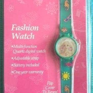 Peut inclure: Un emballage rose et vert pour une montre Barbie pour filles. La montre a un couvercle rabattable pour r&eacute;v&eacute;ler l'heure. Le cadran de la montre a une image d'une poup&eacute;e Barbie. Le bracelet de la montre a un hippocampe, des lunettes de soleil et un motif soleil.