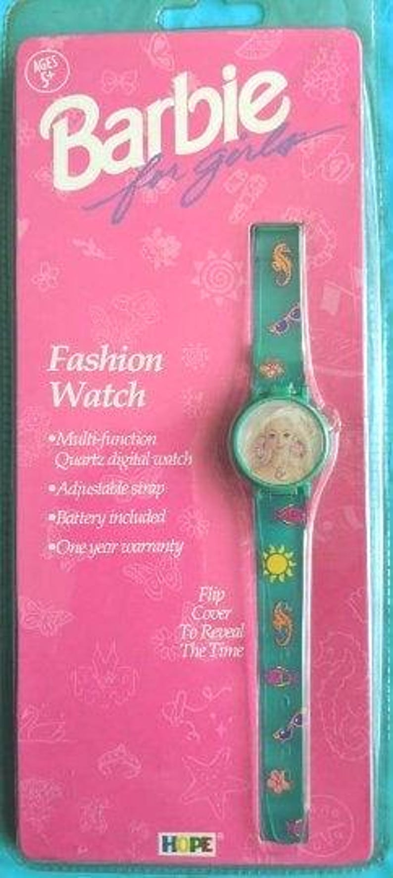 Peut inclure: Un emballage rose et vert pour une montre Barbie pour filles. La montre a un couvercle rabattable pour r&eacute;v&eacute;ler l'heure. Le cadran de la montre a une image d'une poup&eacute;e Barbie. Le bracelet de la montre a un hippocampe, des lunettes de soleil et un motif soleil.