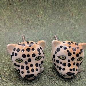 May include: Two ceramic leopard head cabinet knobs. The knobs have a tan base color with black spots, green eyes, and a screw post. The knobs are on a green and brown surface.
