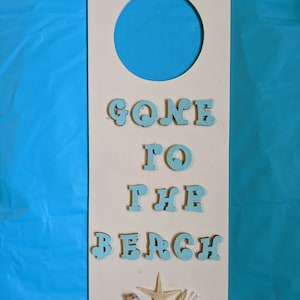 May include: White wooden door hanger with a blue circle cutout. The words "Gone to the Beach" are spelled out in blue letters. There are two small seashells and a starfish on the bottom.
