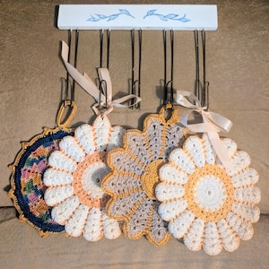 May include: A collection of handmade crochet trivets in various colors, including white, peach, yellow, and navy blue. Each trivet is flower-shaped and hangs from a white wooden rack with a floral design.