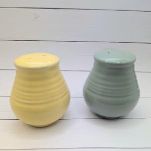 May include: A pair of ceramic salt and pepper shakers. One is yellow, the other is a muted green. Both have a ribbed design and small holes on top. The shakers are on a white wooden surface.