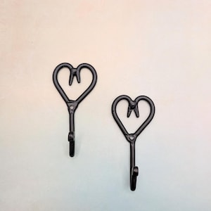May include: Two black metal hooks shaped like hearts with a tooth design inside. The hooks are mounted on a white wall.