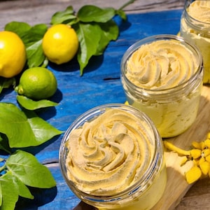 May include: Three glass jars filled with a yellow whipped cream-like substance. The jars are on a wooden surface with lemons and limes in the background.