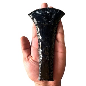 May include: A black obsidian arrowhead with a pointed tip and a wide, flared base. The arrowhead is held in a hand, showcasing its size and detail.